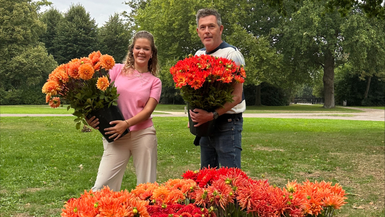 Twee mensen houden grote potten met oranje bloemen vast in een parkachtige omgeving, omringd door bomen en gras.