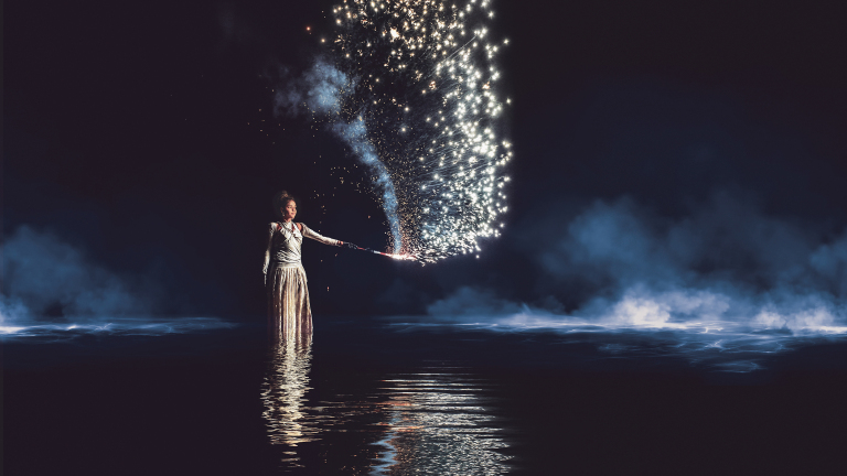 Een vrouw in een jurk staat in een donkere omgeving met water, terwijl ze een stok met vuurwerk in haar hand houdt die vonken en licht verspreidt.
