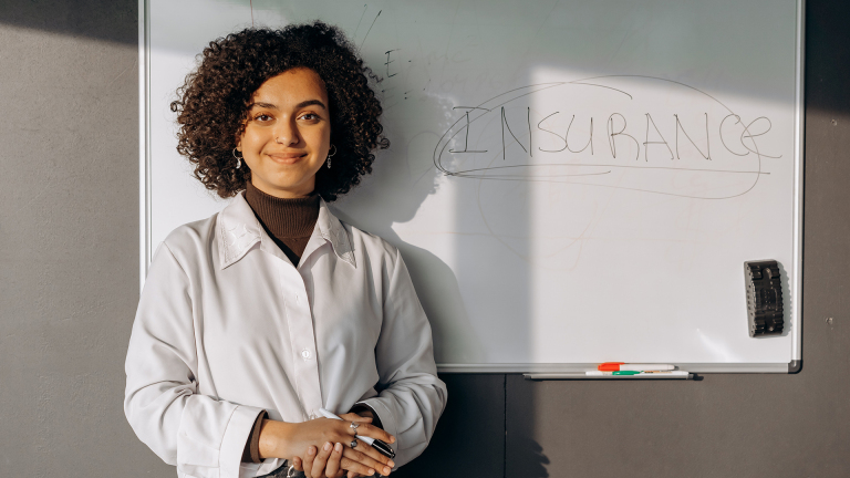 Een vrouw met krullend haar staat voor een whiteboard waarop het woord 'INSURANCE' is geschreven, ze draagt een wit overhemd en houdt pennen vast.