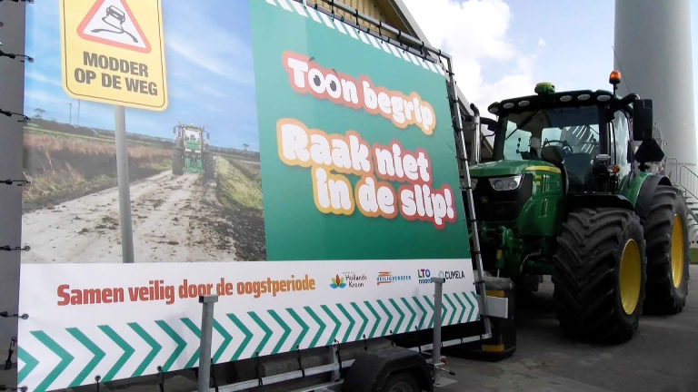 Grote tractor naast een bord met de tekst 'Toon begrip, raak niet in de slip!' en een afbeelding van een modderige weg.