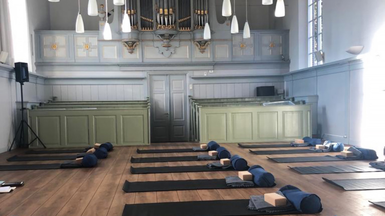 Binnenkant van een gebouw met yoga matten en kussens op de vloer, klaar voor een yogasessie. Het interieur heeft grijs-groene houten panelen en een groot pijporgel aan de muur.
