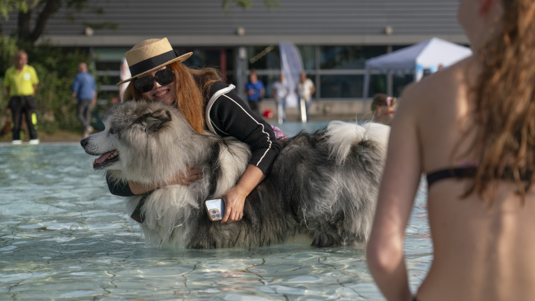 Vrouw met zonnehoed en zonnebril knuffelt een grote pluizige hond in een zwembad, terwijl een andere vrouw op de voorgrond staat.