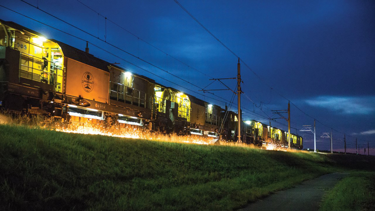 Een trein rijdt 's nachts, verlicht door gele lichten met vonken die van de rails springen onder een blauwe hemel.