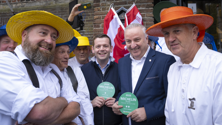 Groep mannen in witte kleding en kleurrijke hoeden poseren met Michelin-gidsen voor een bakstenen gebouw, terwijl een persoon foto's maakt.
