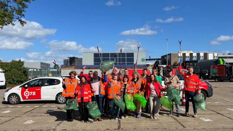 Een groep mensen in oranje veiligheidshesjes poseert op een parkeerplaats met grijpers en groene afvalzakken, klaar voor een schoonmaakactie.