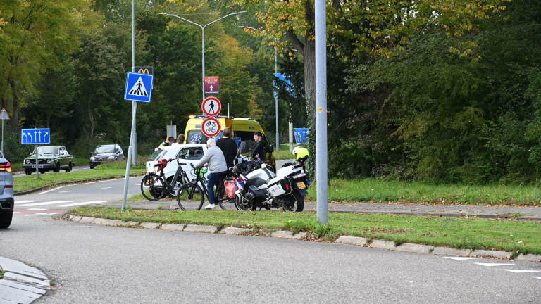 Verkeersincident langs de weg met een ambulance, politie, en een groep mensen bij een scooter en fiets in een groene, beboste omgeving.