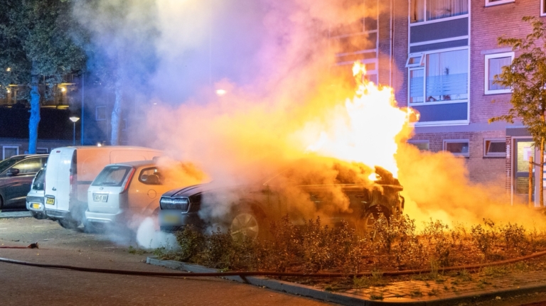 Auto in brand op een parkeerplaats in een woonwijk, met vlammen en rookwolken opstijgend nabij geparkeerde voertuigen en een woongebouw.