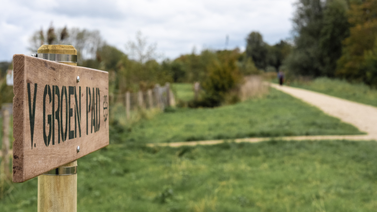 Houten bord met de tekst 'Groen Pad' langs een landelijk wandelpad omgeven door gras en bomen op een bewolkte dag.