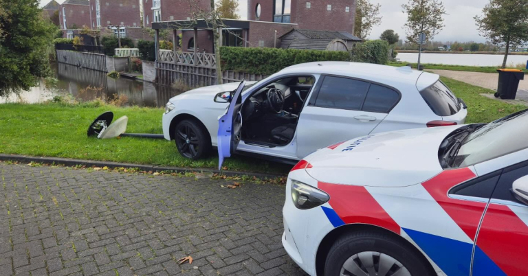 Een wit voertuig met geopende voordeur is naast een omgevallen verkeersbord geparkeerd op het gras, met een politieauto ervoor geparkeerd. Op de achtergrond zijn water en huizen zichtbaar.