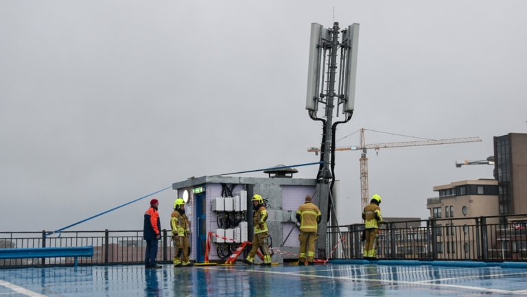 Brandweerlieden inspecteren een antennemast op het dak van een gebouw op een regenachtige dag.