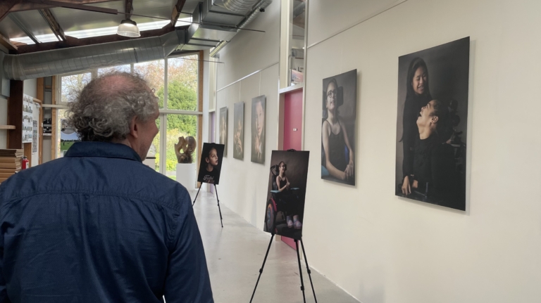 Een man bekijkt portretten die langs een lichte muur in een galerij zijn opgehangen, met enkele schilderijen op ezels, waaronder een portret van een meisje in een rolstoel.