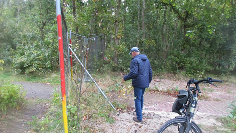 Een man in een blauwe jas en pet loopt in de richting van een hek in een bosrijke omgeving, met een fiets op de voorgrond.