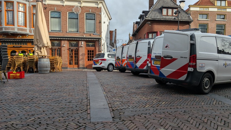 Parkeerplaats in een geplaveide straat met drie politievoertuigen en een café op de achtergrond, met opgestapelde stoelen en tafels onder een grote parasol.