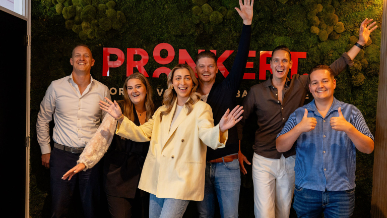 Een groep van zes mensen staat lachend voor een moswand bij een bord met de tekst 'PRONKERT'.