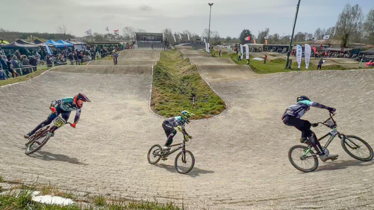 Drie BMX-fietsers nemen een bocht op een verharde racebaan tijdens een wedstrijd, omgeven door toeschouwers en tenten op de achtergrond.