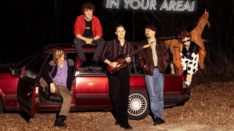 Vijf mensen poseren rond een rode auto; een van hen houdt een gitaar vast, anderen lachen ontspannen.