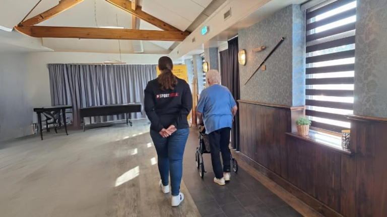 Twee mensen lopen door een lichte kamer; een oudere persoon met een rollator en een jongere persoon in een sportjas.