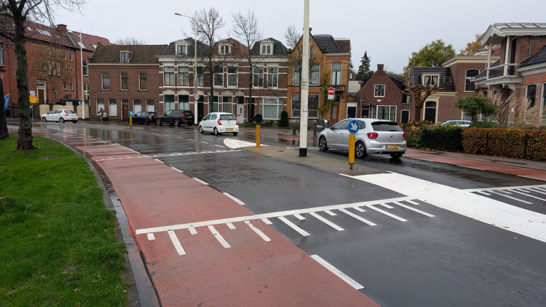 Straat in een woonwijk met bakstenen huizen, regenachtige omstandigheden, en een rood fietspad naast de weg. Auto’s zijn geparkeerd langs de straat, en er rijdt een auto op de rijbaan. Verkeersborden en markeringen zijn zichtbaar op de natte weg.