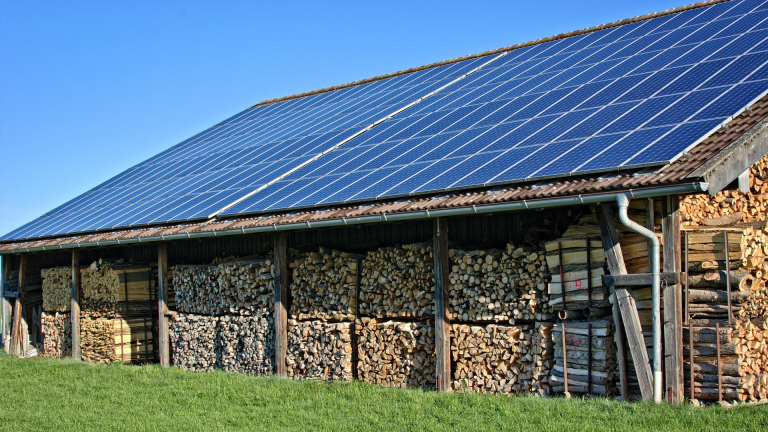 Opslagplaats voor hout met zonnepanelen op het dak, gelegen op een grasveld onder een blauwe hemel.