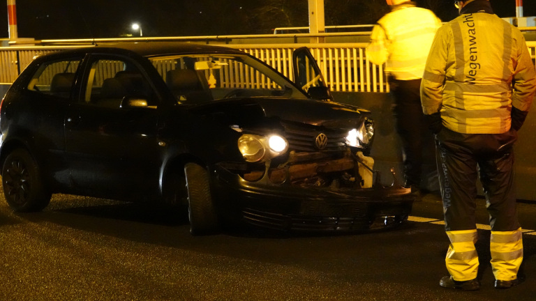 Een beschadigde zwarte auto op een weg met twee wegenwachten die ernaast staan.