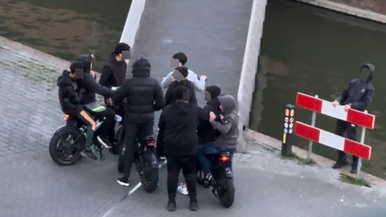 Een groep mensen verzameld rond elektrische fietsen bij een kanaal, met een persoon op de achtergrond bij een verkeersbarrière.