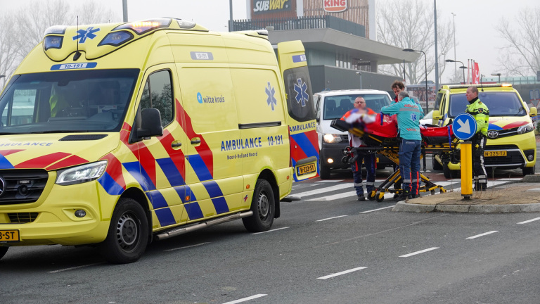 Ambulancepersoneel vervoert een persoon op een brancard naar een ambulance bij een drukke weg nabij winkels.