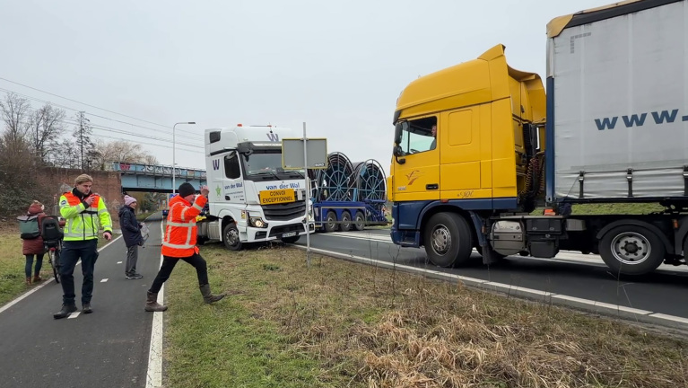 Vrachtwagens op de snelweg met verkeersbegeleiders in oranje jassen die het verkeer regelen; mensen staan langs de weg.