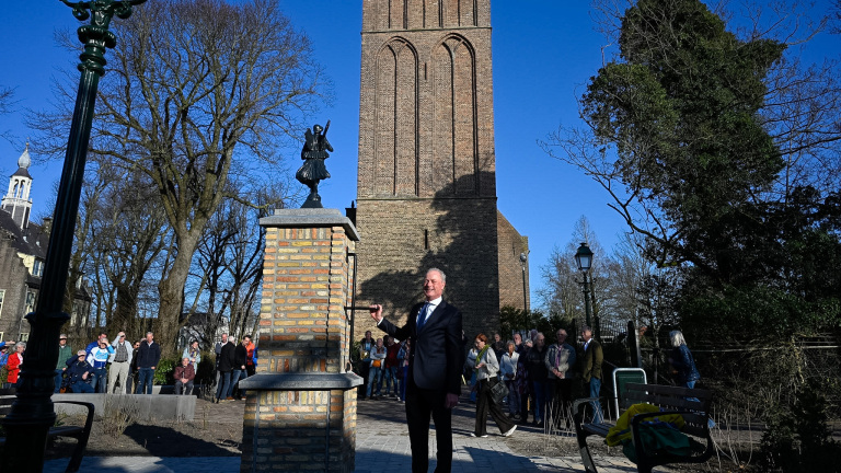 Man onthult een standbeeld op een bakstenen sokkel met een toren op de achtergrond en een groep toeschouwers.