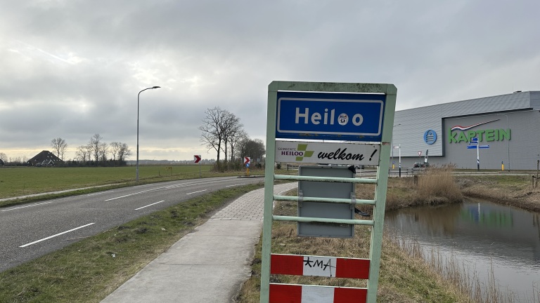 Welkomstbord van Heiloo naast een weg met grasvelden en een groot gebouw op de achtergrond.