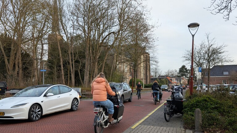 Fietsers en auto's op een smalle weg omgeven door bomen en gebouwen op een bewolkte dag.