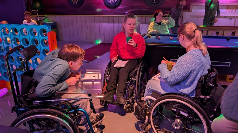 Mensen in rolstoelen houden een gezellige bijeenkomst in een ruimte met een pooltafel en bordspellen.