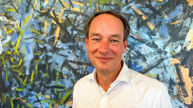 Man in wit overhemd staat voor een kleurrijk abstract schilderij met blauwe en gele tinten.