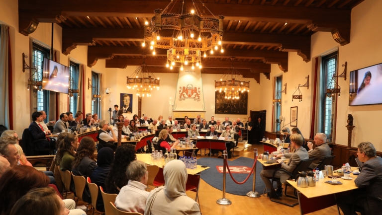 Vergaderzaal vol mensen zittend in een kring, met decoratieve kroonluchters en grote schermen aan de muren.