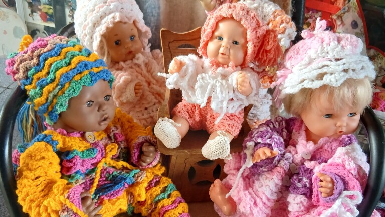 Vier poppen in kleurrijke gehaakte kleding, waarvan één in een houten stoel zit.