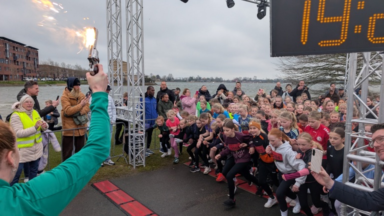 Een grote groep kinderen start een race na het afvuren van een startpistool, met toeschouwers langs de kant.
