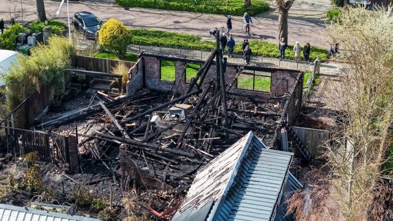 Verwoest gebouw door brand, met verkoolde houten balken en muren. Kijkers verzamelen zich langs de straat.