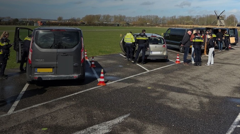 Politieagenten controleren voertuigen op een parkeerplaats met enkele mensen in gesprek, met een windmolen op de achtergrond.