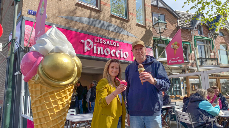 Twee mensen staan voor een ijssalon genaamd Pinoccio, elk met een ijsje in de hand, naast een groot beeld van een ijshoorntje. Het is een zonnige dag en er zijn mensen op het terras te zien.