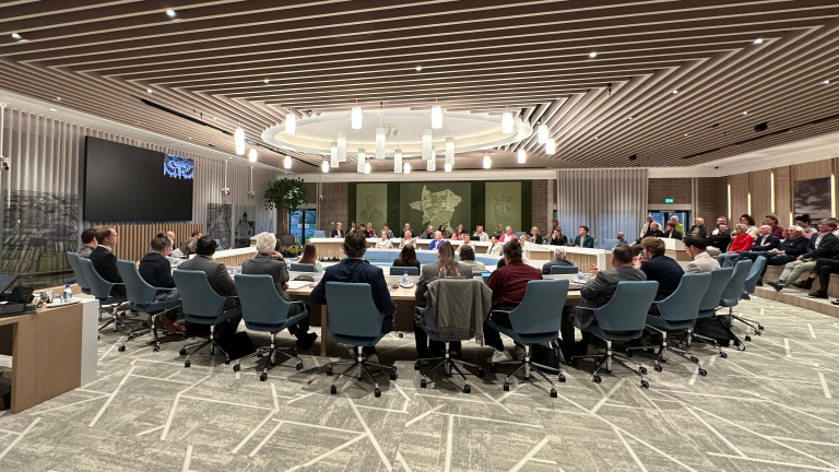 Een groep mensen zit in een moderne vergaderzaal rond een grote ovale tafel met blauwe stoelen.