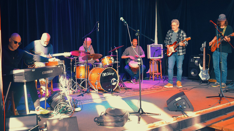 Een band van zes mannen speelt verschillende instrumenten op een podium met een donkere achtergrond.
