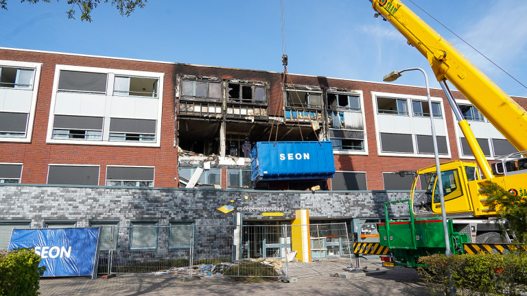 Een beschadigd gebouw met zichtbare roetschade, waar een grote gele hijskraan een blauwe container van SEON omhoog tilt.