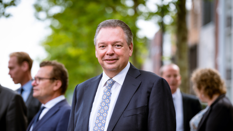 Man in pak met stropdas, staand buiten met onscherpe mensen op de achtergrond.