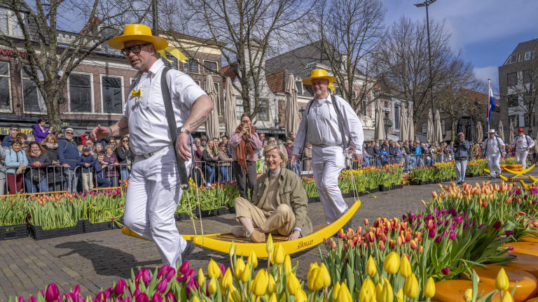 Mannen in witte pakken en gele hoeden dragen een vrouw op een kaasdrager, omringd door kleurrijke tulpen en toeschouwers.