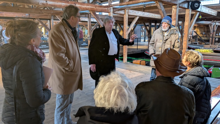 Een groep mensen luistert naar een spreker in een houten gebouw met veel balken.