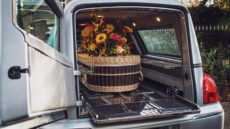 Gevlochten lijkkist met bloemen in de open achterkant van een zilveren begrafenisauto.