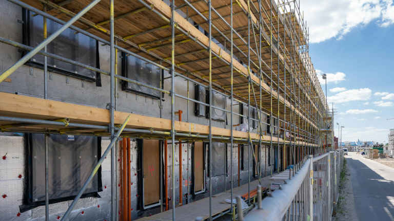 Nieuwbouwproject met steigers langs een onafgewerkt gebouw onder een zonnige blauwe hemel.