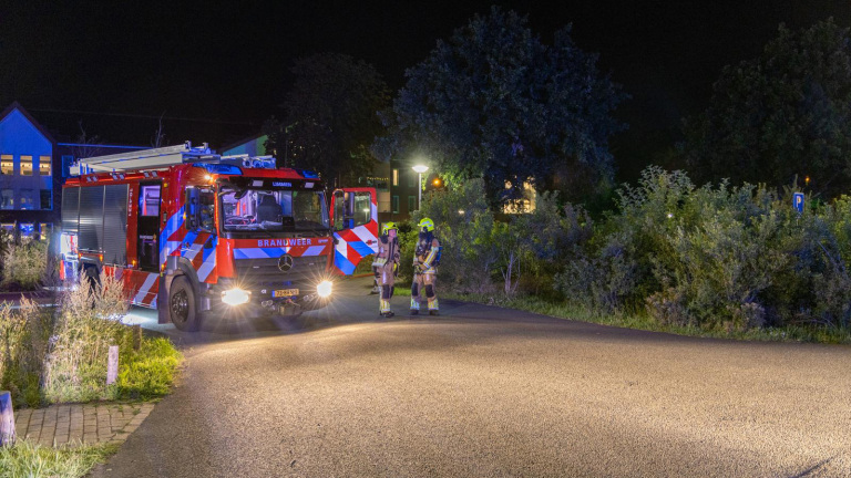 Brandweerwagen met zwaailichten aan, geparkeerd op een weg bij nacht met twee brandweerlieden in uniform ernaast; bomen en een gebouw op de achtergrond.