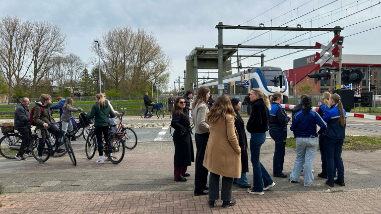 Mensen wachten bij een gesloten spoorwegovergang terwijl een trein passeert, met enkele voetgangers en fietsers.