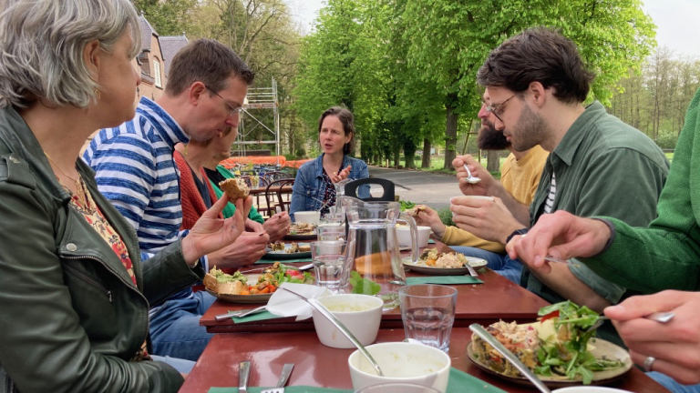 Groep mensen eet samen aan een lange tafel buiten, omringd door groene bomen.