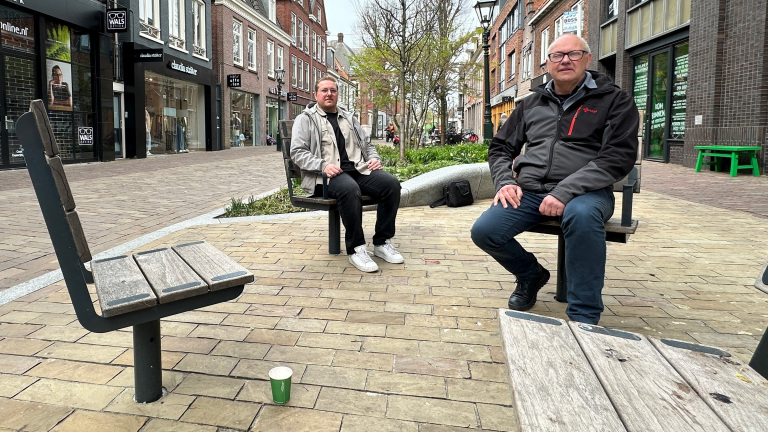 Twee mannen zitten op tegenover elkaar geplaatste banken in een winkelstraat met logo's van opticiens zichtbaar op de achtergrond.